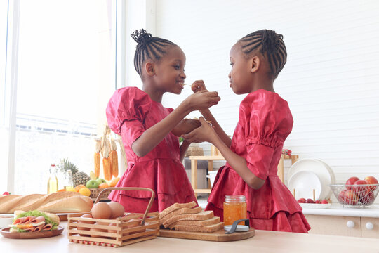 African twin girl sister with curly hair braid African hairstyle eating snack chips in kitchen. Happy smiling kid sibling spending time together. Cute children in lovely family.