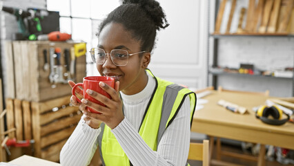 A smiling black woman wearing safety glasses and a reflective vest holds a red mug in a well-equipped carpentry workshop.