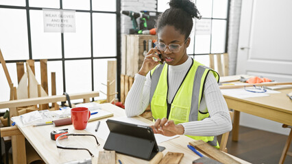 African american woman carpenter in safety vest using phone and tablet in woodwork workshop