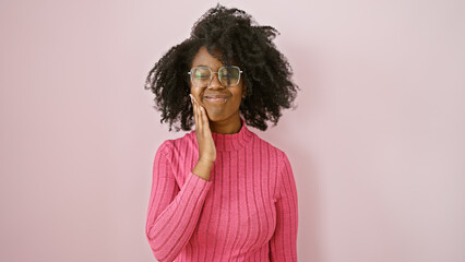 A smiling african american woman in pink holds her cheek, suggesting toothache, against a plain...