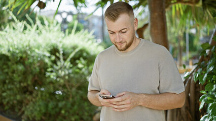 Bearded young man using smartphone in sunny park surrounded by greenery