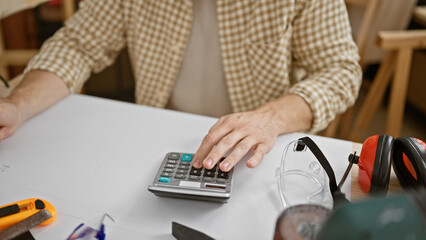 A man calculates expenses in his carpentry workshop, surrounded by tools and safety gear.