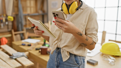 A young woman with tattoos checks her phone in a well-equipped carpentry studio, indicating professional woodworking. - obrazy, fototapety, plakaty