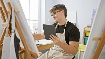 Handsome, confident young hispanic artist smiling, mastering his drawing skills on touchpad at modern art studio indoors