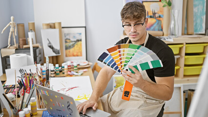 Attractive young hispanic man artist, choosing vibrant paint color on laptop in buzzing art studio