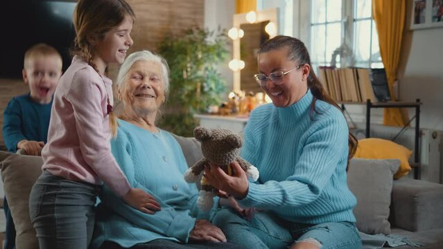 Little girl gives her old grandmother a stuffed toy, multi-generational family spending time together at home, multi generation family sitting together on the sofa at home