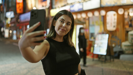 Confident, beautiful hispanic woman smiles, taking a selfie on tokyo street, enjoying city lights at night, capturing the fun through her smartphone.