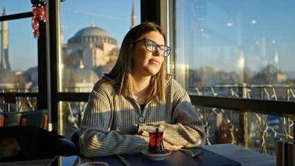 A contemplative young woman sips tea in a turkish restaurant with the hagia sophia in the background.