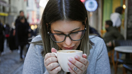 A young woman wearing glasses enjoys a cup of coffee at an outdoor cafe terrace surrounded by urban...