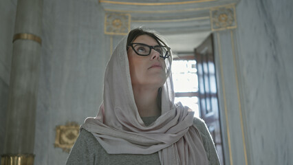 A thoughtful woman wearing glasses and a hijab gazes upwards inside an ornate istanbul mosque.