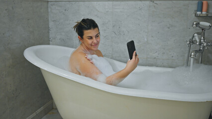 Smiling woman enjoys a bubble bath while using a smartphone in a tiled bathroom