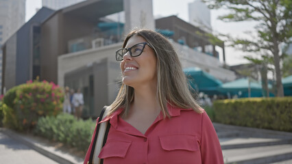 Obraz premium A smiling young adult hispanic woman with brunette hair wearing glasses in a dubai city marina setting.