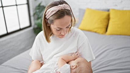 Devoted mother sitting on a relaxed bedroom bed indoors, showing utmost concentration while lovingly breastfeeding her infant. cherishing the athletic home-nurturing lifestyle with seriousness. - obrazy, fototapety, plakaty