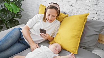 Exhausted mother lovingly typing a message on her smartphone, while her innocent baby daughter...