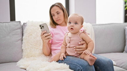 Relaxed mother, sitting on the sofa, having a serious expression as she texts on her smartphone, sharing a lovely moment with her infant daughter at home - obrazy, fototapety, plakaty
