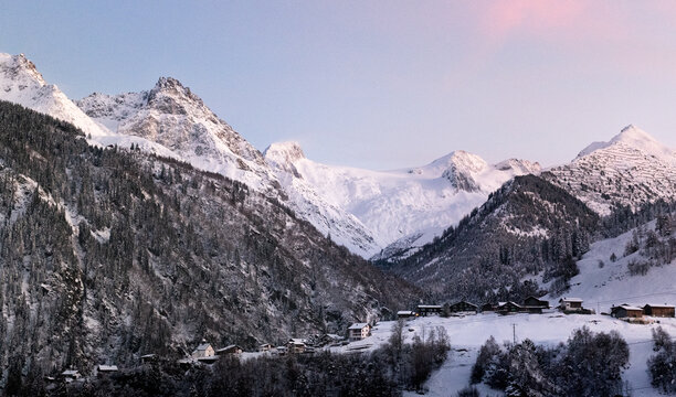 Winter magic in the Swiss Alps: A village, snowy forest, and pink-hued peaks under the evening sun. A grand glacier adds to the picturesque panorama.