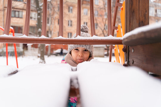 Curious child peeking through snow-covered playground equipment in winter attire