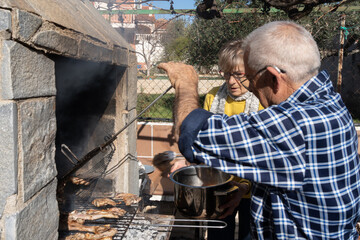 86-year-old man grilling meat on barbecue, dressed in checked shirt, glasses and hearing aid in ear.  73-year-old woman helping to remove meat from the grill, in the background in a mustard yellow jum