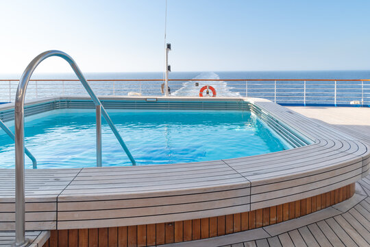 A swimming pool with clear, clean water at the stern of a cruise ship with a view of the ship's wake stretching to the horizon. Wooden deck, handrails and lifebuoy.