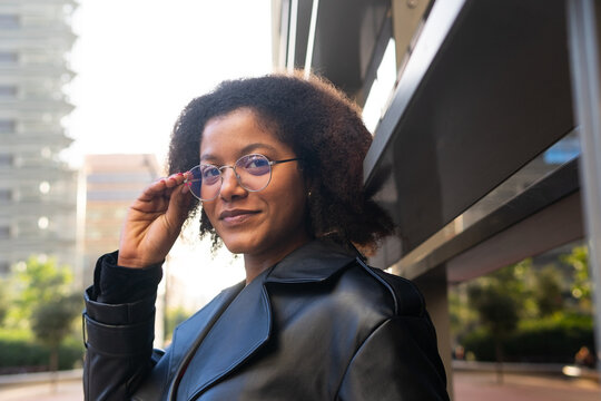 Confident woman adjusting her glasses, dressed in a trendy black coat, with modern architecture in the background