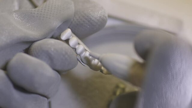 Dental technician polishing artificial ceramic teeth using sandblaster in the dental lab. Professional wearing gloves while holding fake teeth in hand and polishing it with sand blaster.