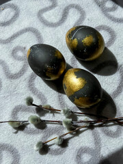 Black and gold eggs lie on a light blanket on the windowsill under the rays of the sun, next to a bunch of willow. Easter Postcard
