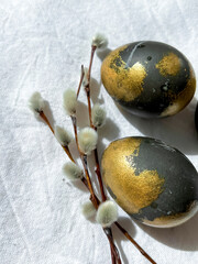Dark blue and gold Easter eggs lie on a white light tablecloth under the rays of the sun, willow branches lie nearby. An Easter card. Vertical photo