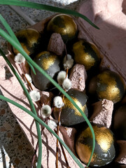 Easter eggs painted in black and gold are lying in a tray next to green leaves of grass. The concept of the spring Easter holiday. Vertical photo
