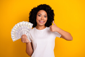 Portrait of satisfied cheerful girl with perming coiffure holding money showing thumb up get salary...