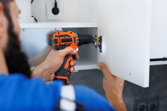 A man in uniform screws the kitchen cabinet doors with an electric screwdriver.