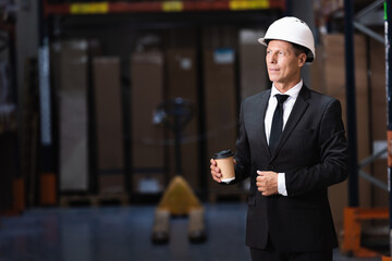 pleased middle aged businessman in hard hat and suit holding coffee in warehouse, professional