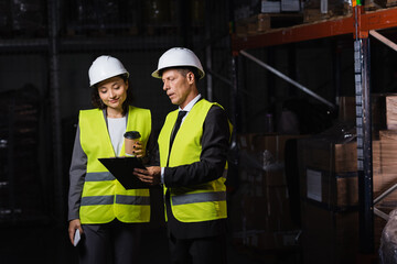 middle aged worker in hard hat showing clipboard to female employee with coffee and smartphone