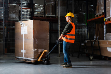 strong middle aged warehouse worker in hard hat and safety vest transporting pallet with hand truck