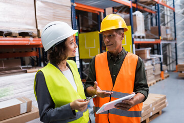 supervisor in hard hat holding tablet while discussing work to happy female employee in warehouse - 740021136
