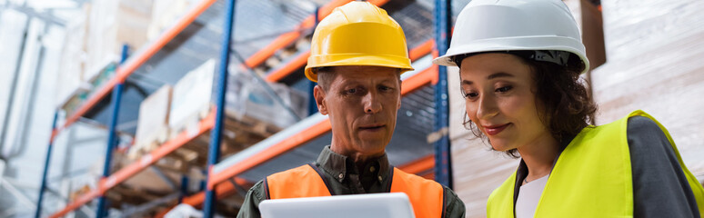 happy supervisor in hard hat holding tablet while explaining work to employee in warehouse, banner