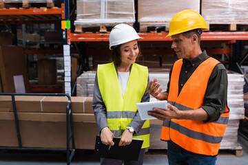 happy supervisor in hard hat holding tablet while explaining work to employee in warehouse - 740021197