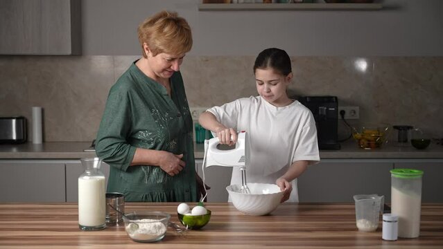 Happy grandmother teach her granddaughter to cook, stirring eggs make dough enjoy process. Share knowledge to younger generation, enjoy communication and friendly relations. International women's day