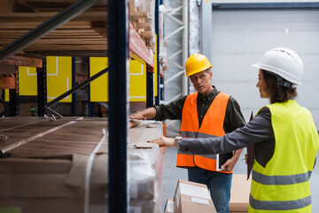 middle aged supervisor in hard hat holding tablet while explaining work to employee in warehouse