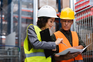 middle aged warehouse supervisor in safety vest showing paperwork to his female employee with pen