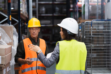 middle aged supervisor in hard hat having conversation with his female employee in warehouse