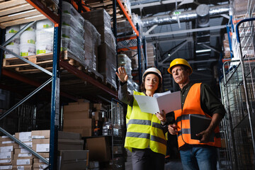 warehouse workers discussing logistics, woman with folder showing direction to middle aged colleague