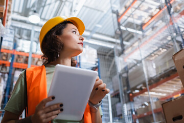 female warehouse worker in safety vest and hard hat holding digital tablet and checking cargo