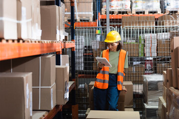 female warehouse worker in safety vest and hard hat holding digital tablet near cargo boxes