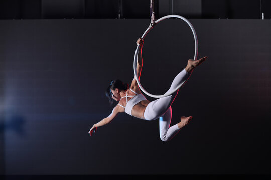 Young woman performing acrobatic element on aerial ring indoors