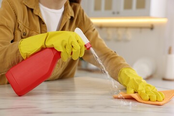 Woman with spray bottle and microfiber cloth cleaning white marble table in kitchen, closeup