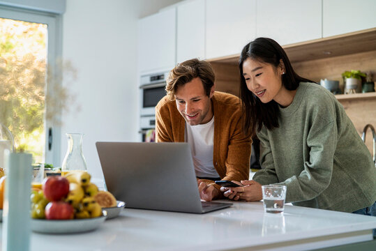 Adult couple shopping online on laptop and smart phone at kitchen counter