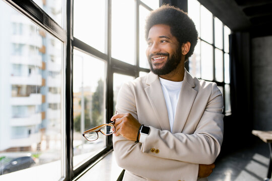 A poised and modern businessman stands by a panoramic window in his office, arms crossed, as he looks out at the city, reflecting the dynamic nature of the modern business world - Powered by Adobe