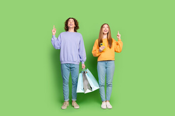 Full length portrait of two astonished people shop bags smart phone look indicate finger up empty...