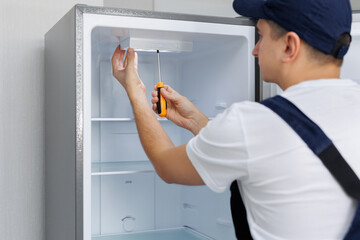 Man in a uniform repairs the light in the refrigerator with a screwdriver. Replacing the light bulb.