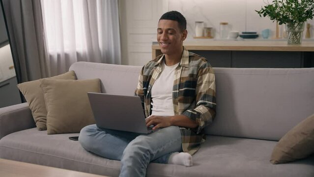 Smiling African American man relaxing at couch apartment biracial ethnic male guy on sofa home mortgage loan user freelancer student businessman using pc laptop computer online shopping pay browsing